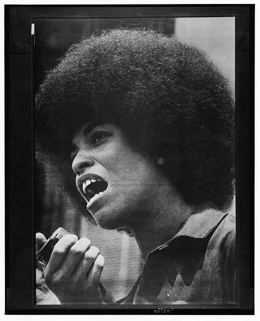 Angela Davis speaking into a microphone, mid-sentence. Close-up black-and-white photograph. Her Afro is full, her expression focused.