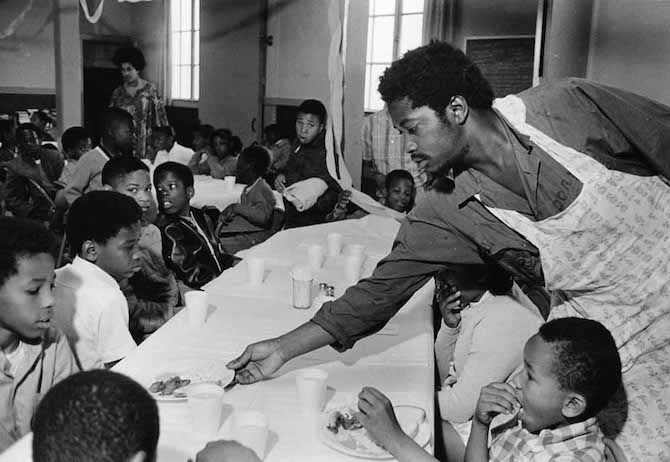 A Black Panther Party member serving breakfast to Black schoolchildren at a long communal table. Multiple children seated and eating, others standing and waiting. Community center interior, 1969.