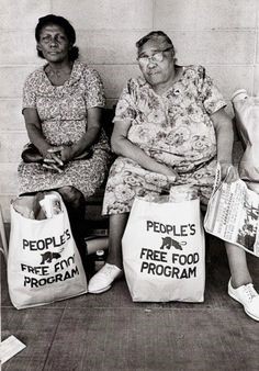 Two elder Black women seated side by side on a stoop, People's Free Food Program paper bags on the ground between them. Both look directly at the camera. Black and white photograph.